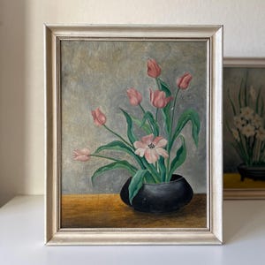 May include: An oil painting in a silver frame shows a bouquet of pink tulips in a black vase. The tulips have green leaves and are set against a grey background. The painting is on a wooden surface.