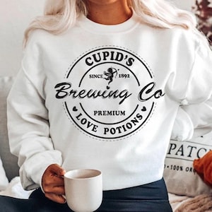 Op de afbeelding: Witte sweatshirt met een zwarte cirkelvormige afbeelding met de tekst "Cupid's Brewing Co", "Since 1892" en "Premium Love Potions". De afbeelding bevat een illustratie van Cupido. Een persoon houdt een witte mok vast.