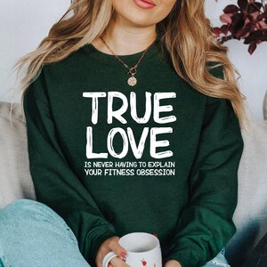 May include: A dark green sweatshirt featuring the words "TRUE LOVE" in large white letters, with the phrase "IS NEVER HAVING TO EXPLAIN YOUR FITNESS OBSESSION" below. The sweatshirt is a solid colour and has long sleeves.
