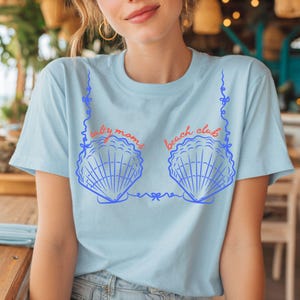 Könnte beinhalten: Ein hellblaues T-Shirt mit einem Grafikdruck von zwei blauen Muscheln, die durch blaue Linien verbunden sind. Der Text "salty moms" ist in Rot über der linken Muschel und "beach club" in Rot über der rechten Muschel aufgedruckt.
