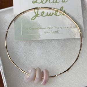 May include: A gold-colored bracelet with four light pink beads. The bracelet is displayed in a white box with a card that reads "Lena's Jewels" and a Bible verse.
