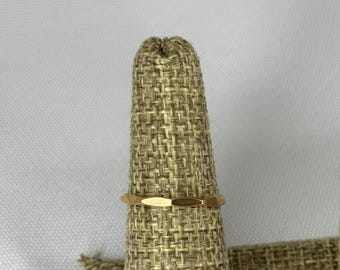May include: A gold-colored ring with a geometric design. The ring is displayed on a textured, tan ring holder. The ring's design features a series of flat, elongated facets around the band.