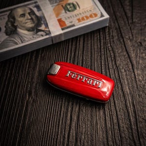 Può includere: Una chiave per auto Ferrari rossa con scritte argentate è appoggiata su una superficie di legno scuro accanto a una pila di banconote da 100 dollari USA. La chiave è il punto focale, con il denaro sullo sfondo.