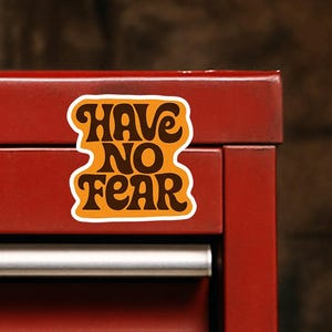 Puede incluir: Una caja de herramientas de metal roja con múltiples cajones y una pegatina que dice "Have No Fear" en una fuente retro. La pegatina es naranja y marrón con un borde blanco. La caja de herramientas tiene tiradores plateados y una cerradura pequeña y redonda.