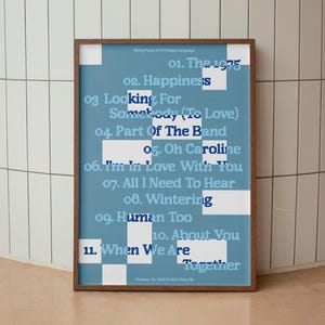 Può includere: Un poster incorniciato con sfondo blu e forme geometriche bianche. Il poster elenca i titoli delle canzoni in un carattere blu scuro, tra cui "The 1975", "Happiness" e "Part of The Band". Il poster è in una cornice di legno marrone.