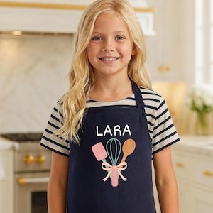 以下が含まれることがあります： 白で「LARA」とプリントされた紺色のエプロン。泡立て器、ヘラ、木のスプーンのグラフィックとピンクのリボンが付いています。縞模様のシャツを着ている人。