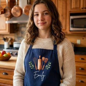 May include: Navy blue apron with a watercolor design of kitchen utensils, including a whisk, rolling pin, and spatula. The name "Emily" is written in cursive script below the design. The apron is worn over a cream-colored sweater.
