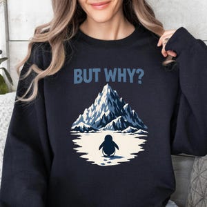 Puede incluir: Sudadera azul marino con un gráfico de pingüino y el texto "BUT WHY?". El gráfico muestra un pingüino frente a una cordillera. El pingüino es blanco y negro, y la montaña es azul y blanca.