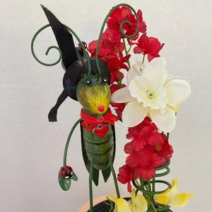 May include: A green grasshopper figurine with a black hat, red bow, and a heart charm. The figurine is standing on a green leaf with a black base. The grasshopper is surrounded by red, white, and yellow flowers.