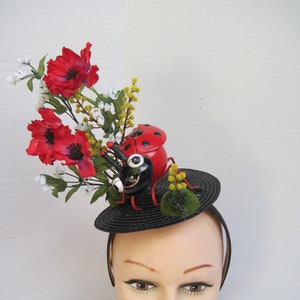 May include: A black headband with a small black hat adorned with a red and black ladybug, green moss, and a cluster of red and white flowers.