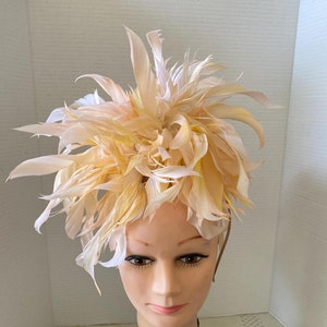May include: A large, fluffy hat made of feathers in shades of peach and cream. The hat is perched on a mannequin head.