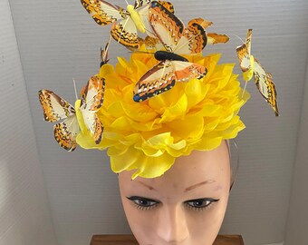 May include: A yellow floral headpiece with several orange and white patterned butterflies attached. The headpiece is on a mannequin head. The butterflies have intricate wing patterns and are positioned around the yellow flower.