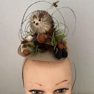 May include: A whimsical hat with a brown hedgehog, a small bird, and acorns on a straw base. The hat is decorated with wire and a bee.