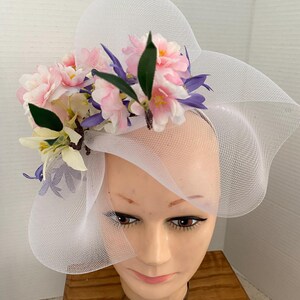 May include: A white mesh hat with a wide brim, adorned with pink, purple, and white artificial flowers and green leaves. The hat is displayed on a mannequin head, showcasing its design and style.