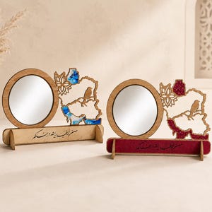 May include: Two wooden desk mirrors with circular glass panes. Each mirror is attached to a map-shaped cutout featuring a bird silhouette and floral patterns. One map has blue resin accents, the other has red glitter. Both have Persian calligraphy.