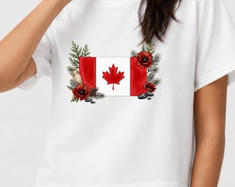 Può includere: Una t-shirt bianca a maniche corte con una grafica centrale della bandiera canadese. La bandiera è incorniciata da illustrazioni in stile acquerello di papaveri rossi, rami di pino verdi, un pezzo di corteccia di betulla e pietre grigie.