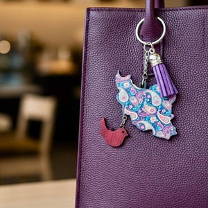 Puede incluir: Un bolso de mano morado oscuro con un asa a juego. Un llavero con un mapa con estampado de cachemira y un pequeño dije de pájaro rojo cuelga del asa. El llavero también tiene una borla morada.