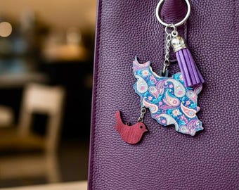 Può includere: Una borsa viola scuro con manico abbinato. Un portachiavi con una mappa a fantasia paisley e un piccolo ciondolo a forma di uccello rosso è appeso al manico. Il portachiavi ha anche un nappa viola.
