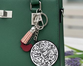 Può includere: Una borsa verde scuro con chiusura e manico argentati. Un portachiavi con nappa rosa, un ciondolo a forma di uccello rosso e un ciondolo rotondo con calligrafia in bianco e nero è attaccato al manico.