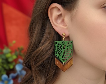 Può includere: Orecchini in legno verdi e marroni con un design a foglie. La parte superiore è verde scuro con un motivo a foglie ritagliate e la parte inferiore è un pezzo marrone a forma di freccia. Gli orecchini pendono da perni color oro.
