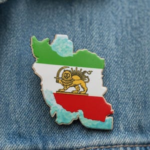Könnte beinhalten: Eine Anstecknadel in Form des Iran, die die Nationalflagge zeigt. Der obere Teil ist grün, die Mitte weiß mit einem goldenen Löwen- und Sonnenemblem und der untere Teil rot. Die Anstecknadel befindet sich vor einem blauen Jeanshintergrund.