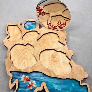 May include: Wooden decorative tray in the shape of a country, featuring a blue resin river with goldfish. An oval wooden piece with red text and a floral design sits atop the tray. The tray has several cut-out sections.