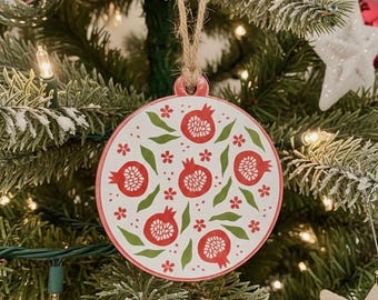 Può includere: Ornamento natalizio rotondo con un design a melograno rosso e bianco. L'ornamento è appeso a un albero di Natale con luci e altre decorazioni. Ha un cordino di iuta per appenderlo.