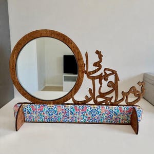 May include: A wooden vanity mirror with a circular frame and a decorative shelf. The shelf is covered in a colourful floral fabric. The mirror has Arabic calligraphy on the top right.