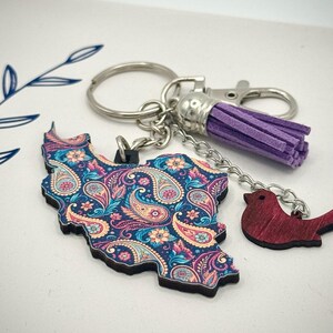 May include: A keychain featuring a paisley-patterned map of a country, with a small red bird charm and a purple tassel. The map is in shades of blue, pink, and yellow. The keychain has a silver clasp and key ring.