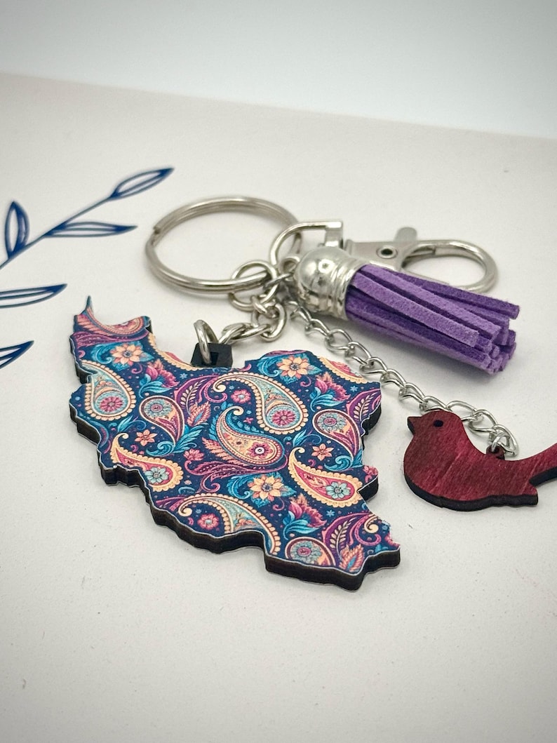 May include: A keychain featuring a paisley-patterned map of a country, with a small red bird charm and a purple tassel. The map is in shades of blue, pink, and yellow. The keychain has a silver clasp and key ring.