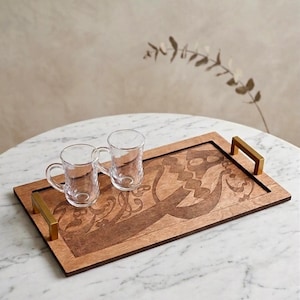 May include: A rectangular wooden serving tray with gold-coloured handles, holding two clear glass teacups. The tray features an engraved design in a darker shade of brown. The tray is on a white marble table.