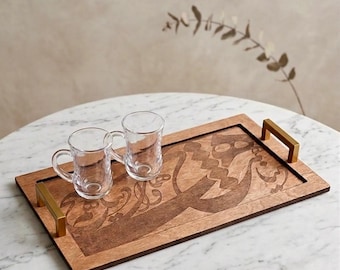 Può includere: Un vassoio rettangolare in legno con manici color oro, che contiene due tazze da tè in vetro trasparente. Il vassoio presenta un disegno inciso in una tonalità più scura di marrone. Il vassoio è su un tavolo di marmo bianco.