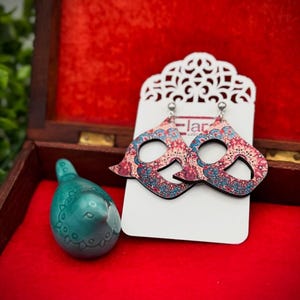 May include: Wooden drop earrings with a paisley pattern in shades of red, blue, and cream are displayed on a white card. A small, teal ceramic bird figurine sits to the left. The background is a red velvet-lined box.