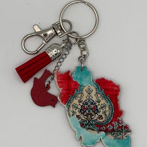 May include: A keychain with a map of Iran in red and blue, a red tassel, and a small red bird charm. It has a silver clasp and a key ring. The map design resembles a Persian carpet.