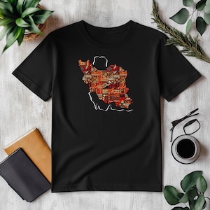Puede incluir: Camiseta negra con un contorno blanco de un mapa de un país lleno de un diseño colorido. El diseño incorpora formas geométricas rojas, naranjas y marrones. La camiseta se muestra sobre una superficie blanca con otros objetos.