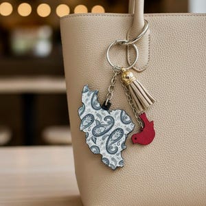 May include: A beige handbag with a silver keyring holding a paisley-patterned charm in the shape of a country and a red bird charm. A beige tassel hangs from the keyring. The bag has a textured surface.