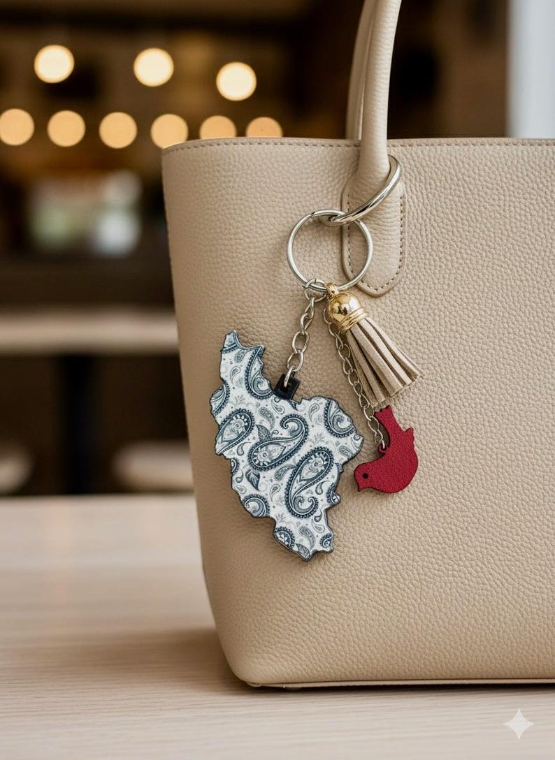 May include: A beige handbag with a silver keyring holding a paisley-patterned charm in the shape of a country and a red bird charm. A beige tassel hangs from the keyring. The bag has a textured surface.