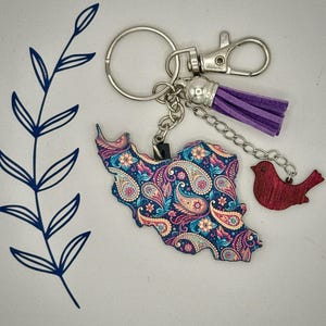 May include: A keychain featuring a paisley-patterned map of Iran in blue, pink, and yellow. It includes a purple tassel, a red bird charm, and silver-tone hardware. The keychain is designed for keys or bags.