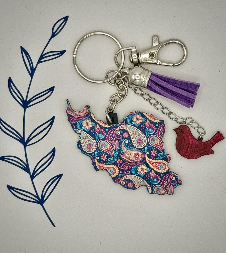 May include: A keychain featuring a paisley-patterned map of Iran in blue, pink, and yellow. It includes a purple tassel, a red bird charm, and silver-tone hardware. The keychain is designed for keys or bags.