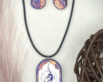 Può includere: Un set di gioielli con collana con pendente e orecchini abbinati. Il pendente e gli orecchini hanno un design floreale blu, rosa e giallo. La collana ha un cordino nero e un pendente ovale con un centro bianco e calligrafia blu.