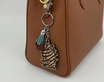 Può includere: Una borsa in pelle marrone con finiture color oro. Un portachiavi decorativo con una nappa turchese e bordeaux e un ciondolo in legno con calligrafia araba è appeso alla borsa.