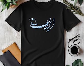 Può includere: T-shirt nero con un disegno blu e bianco di una mappa e una scritta. La grafica è centrata sul davanti. La maglietta è esposta su una superficie bianca con una tazza di caffè, occhiali e altri oggetti.