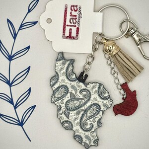 May include: A keychain featuring a paisley-patterned charm in the shape of a state. The charm is white and gray with a black outline. It includes a gold tassel, a red charm, and a tag with the text "Flara design".