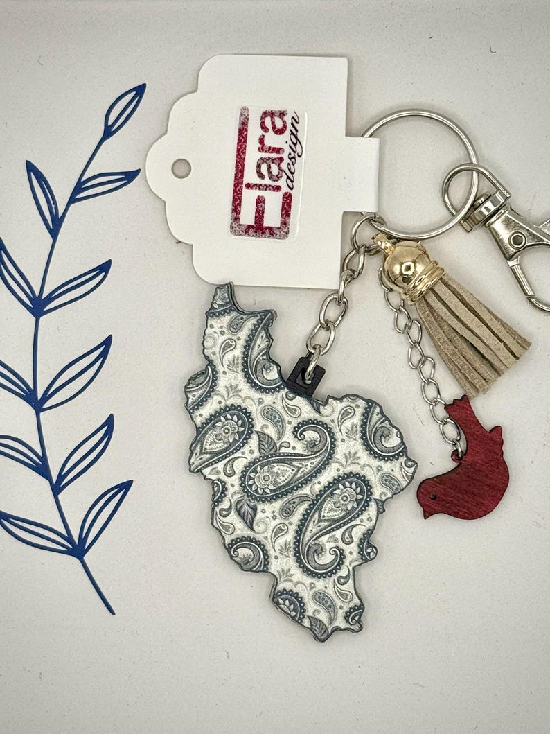 May include: A keychain featuring a paisley-patterned charm in the shape of a state. The charm is white and gray with a black outline. It includes a gold tassel, a red charm, and a tag with the text "Flara design".