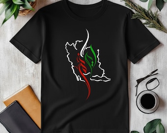 Può includere: T-shirt nero con un contorno bianco di una mappa di un paese e un design rosso, verde e bianco. Il design sembra essere calligrafia o testo stilizzato. La maglietta è mostrata su una superficie chiara.