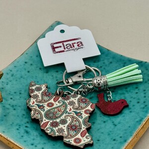 May include: A keychain featuring a paisley-patterned wooden cutout in the shape of a country, with a small red bird charm and a mint green tassel. The keychain includes a silver clasp and a tag that reads "Elara design."