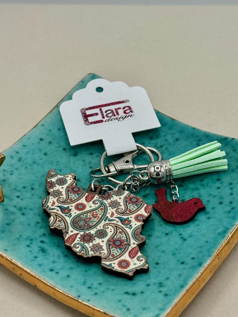 May include: A keychain featuring a paisley-patterned wooden cutout in the shape of a country, with a small red bird charm and a mint green tassel. The keychain includes a silver clasp and a tag that reads "Elara design."