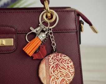 Può includere: Una borsa bordeaux con chiusura e manico dorati è adornata da un portachiavi. Il portachiavi presenta un ciondolo rotondo in legno con calligrafia, un piccolo uccello di legno e un nappa arancione.
