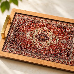 May include: A rectangular wooden serving tray featuring a central inlay with a detailed red, cream, and black floral pattern reminiscent of a woven rug. The tray includes two brushed gold metal handles attached to the sides of the light wood frame.