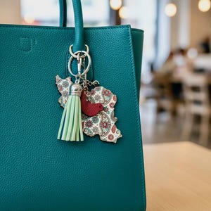 May include: Teal leather handbag with a decorative keychain. The keychain features a light green tassel, a red bird, and a wooden cutout in the shape of a country with a paisley design. The bag has a textured surface.